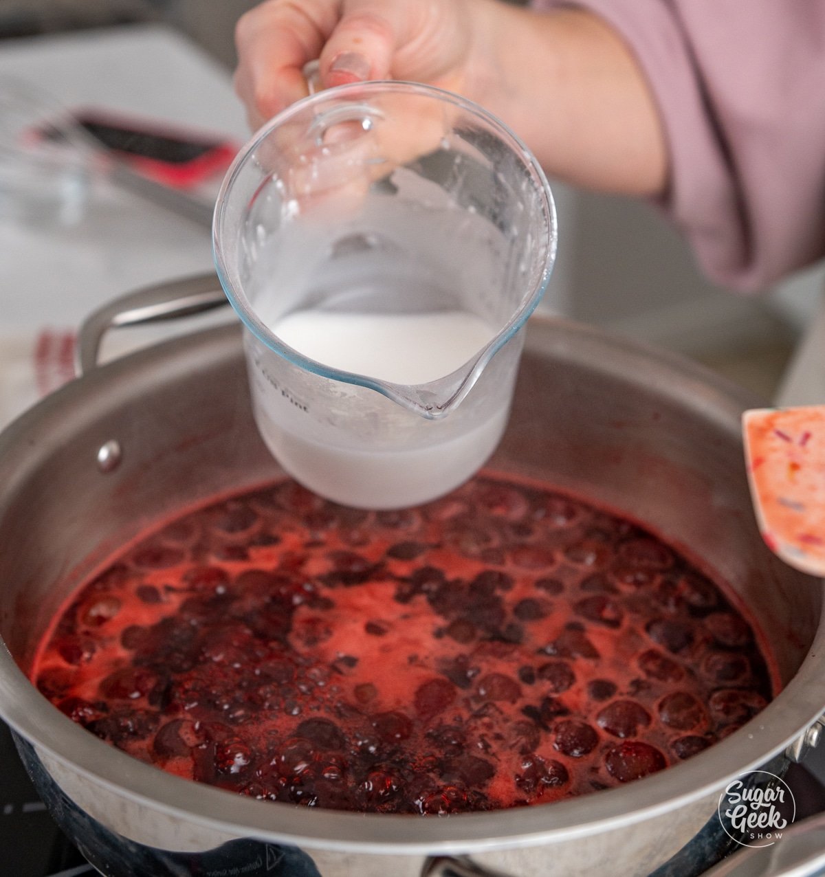 hand adding a measuring cup of cornstarch slurry to cherry filling