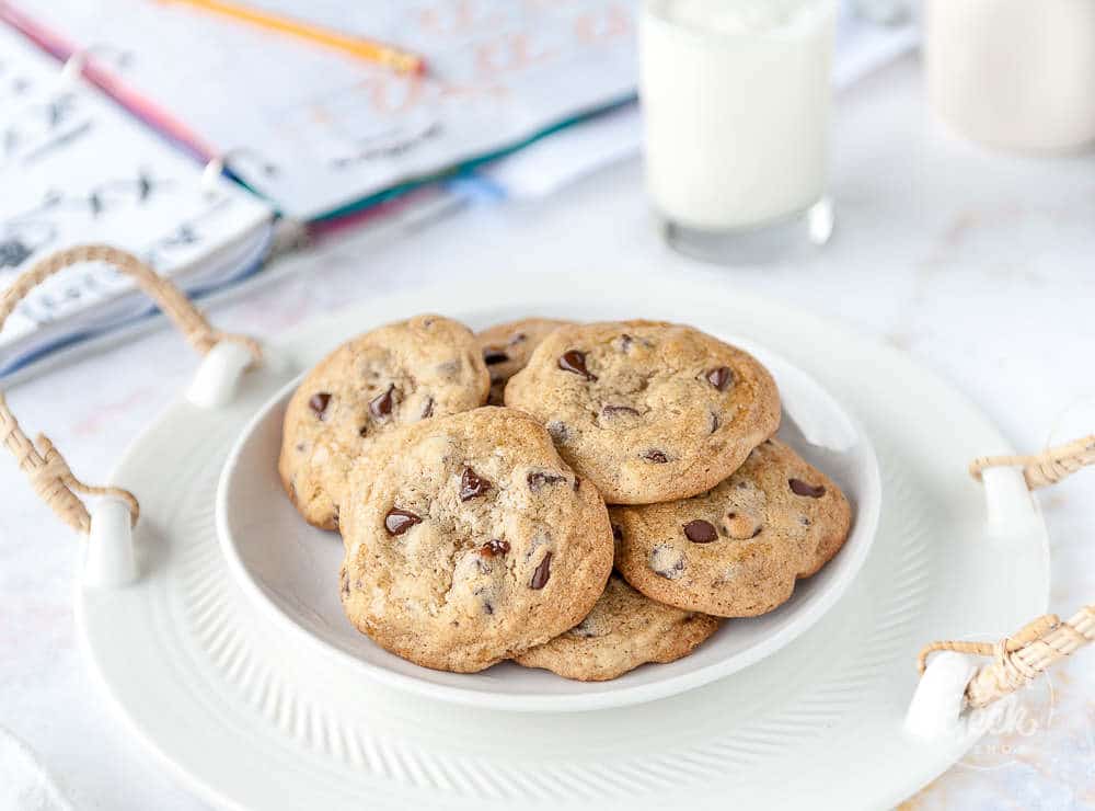 plate of chocolate chip cookies in a white bowl with. schoolbook and glass of milk behind it