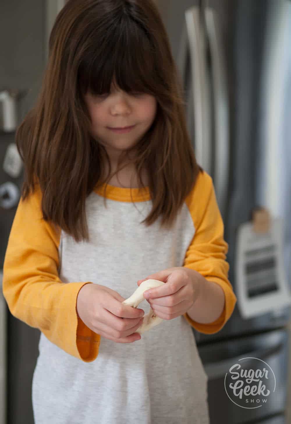 Little girl with brown shoulder-length hair and white and yellow shirt shaping a bagel with her hands