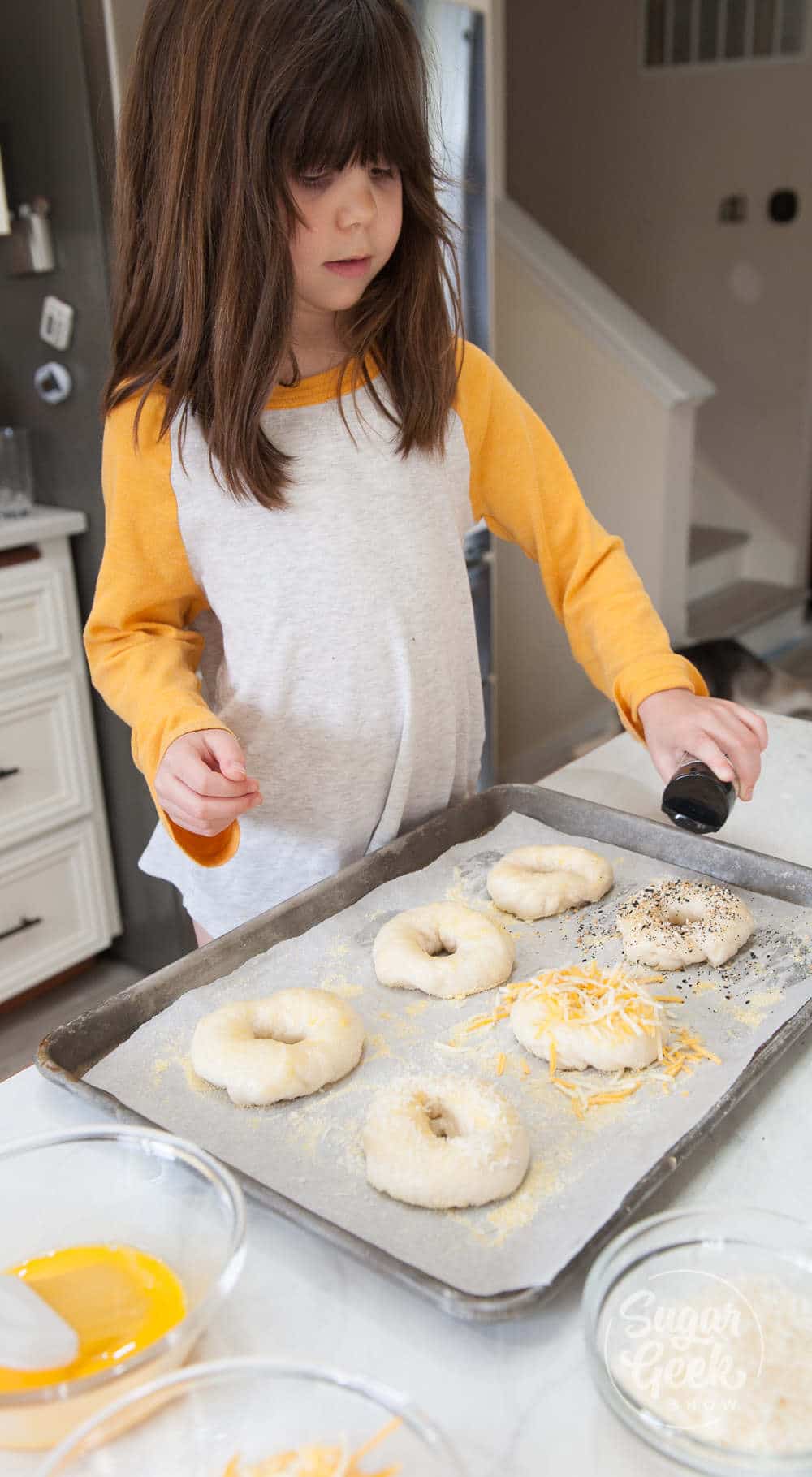 little girl with shoulder-length brown hair and yellow and white shirt sprinkling toppings onto bagels on a sheet pan