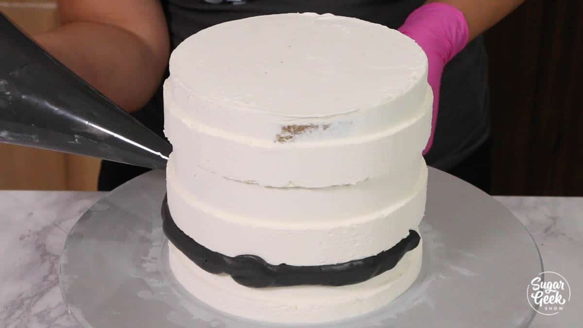 adding black buttercream stripes to white buttercream cake with a piping bag