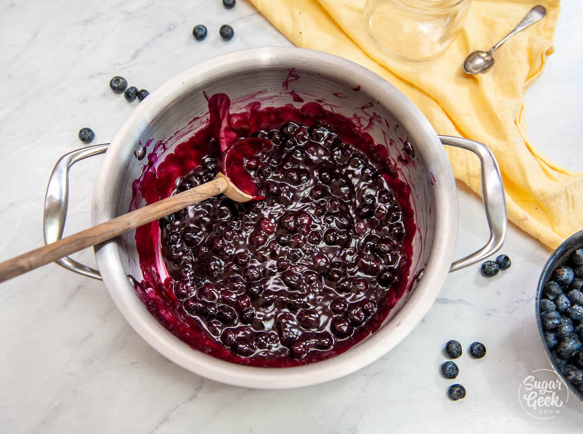 overhead shot of blueberry filling in a stainless steel pot with a wooden spoon and yellow napkin to the side