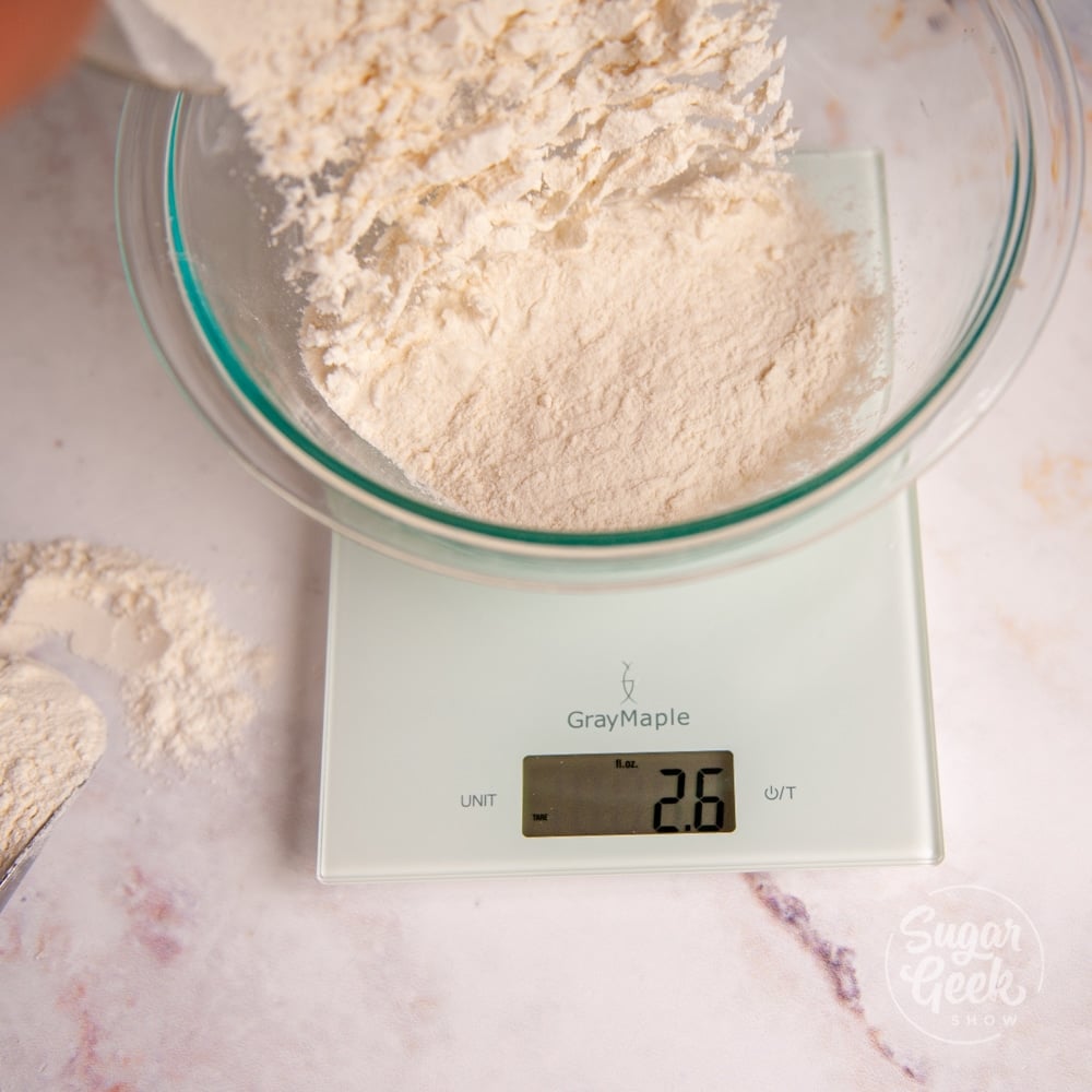 flour being added to a clear mixing bowl on top of a digital kitchen scale