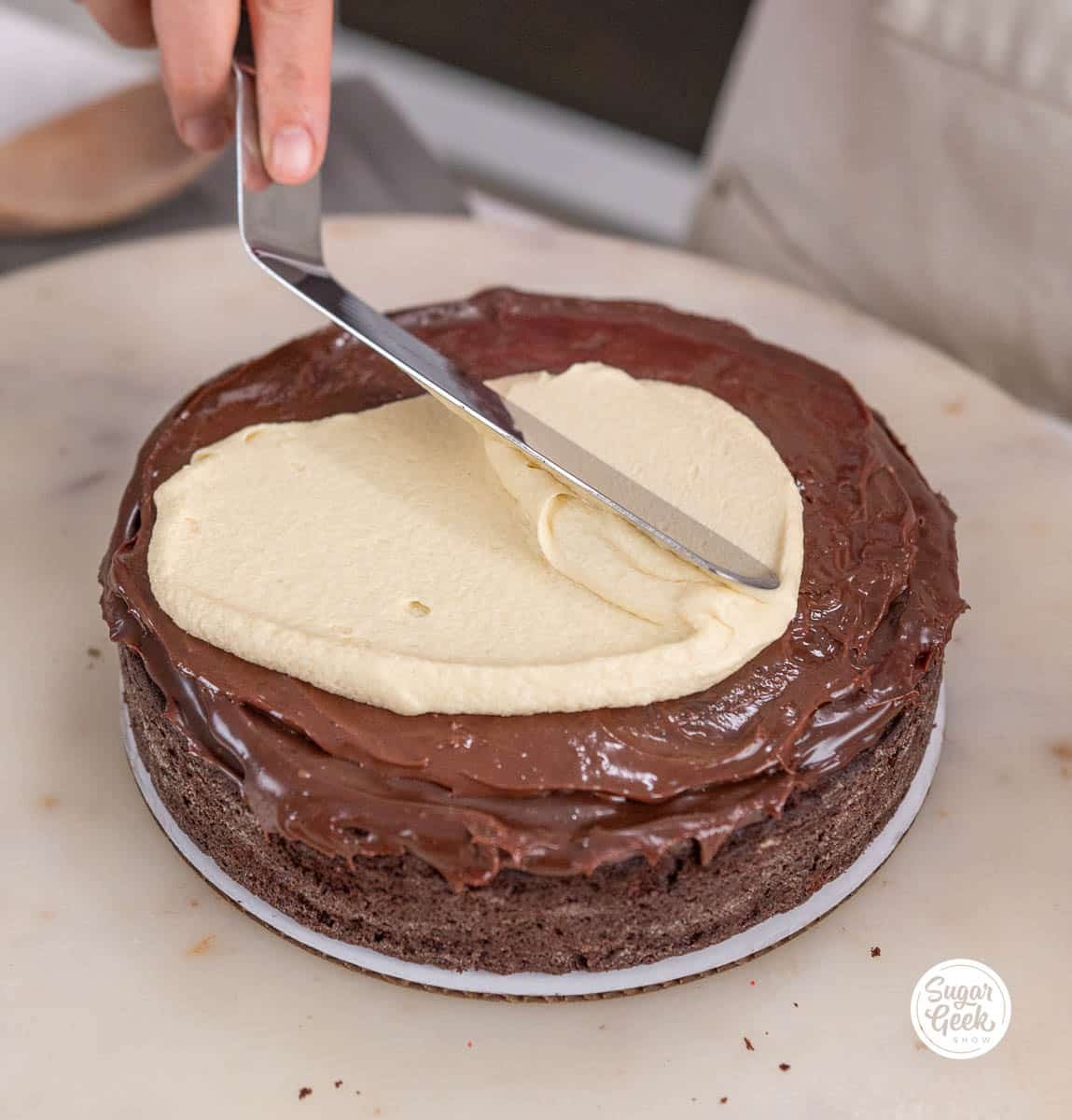 hand smoothing white chocolate ganache with a spatula onto a chocolate cake