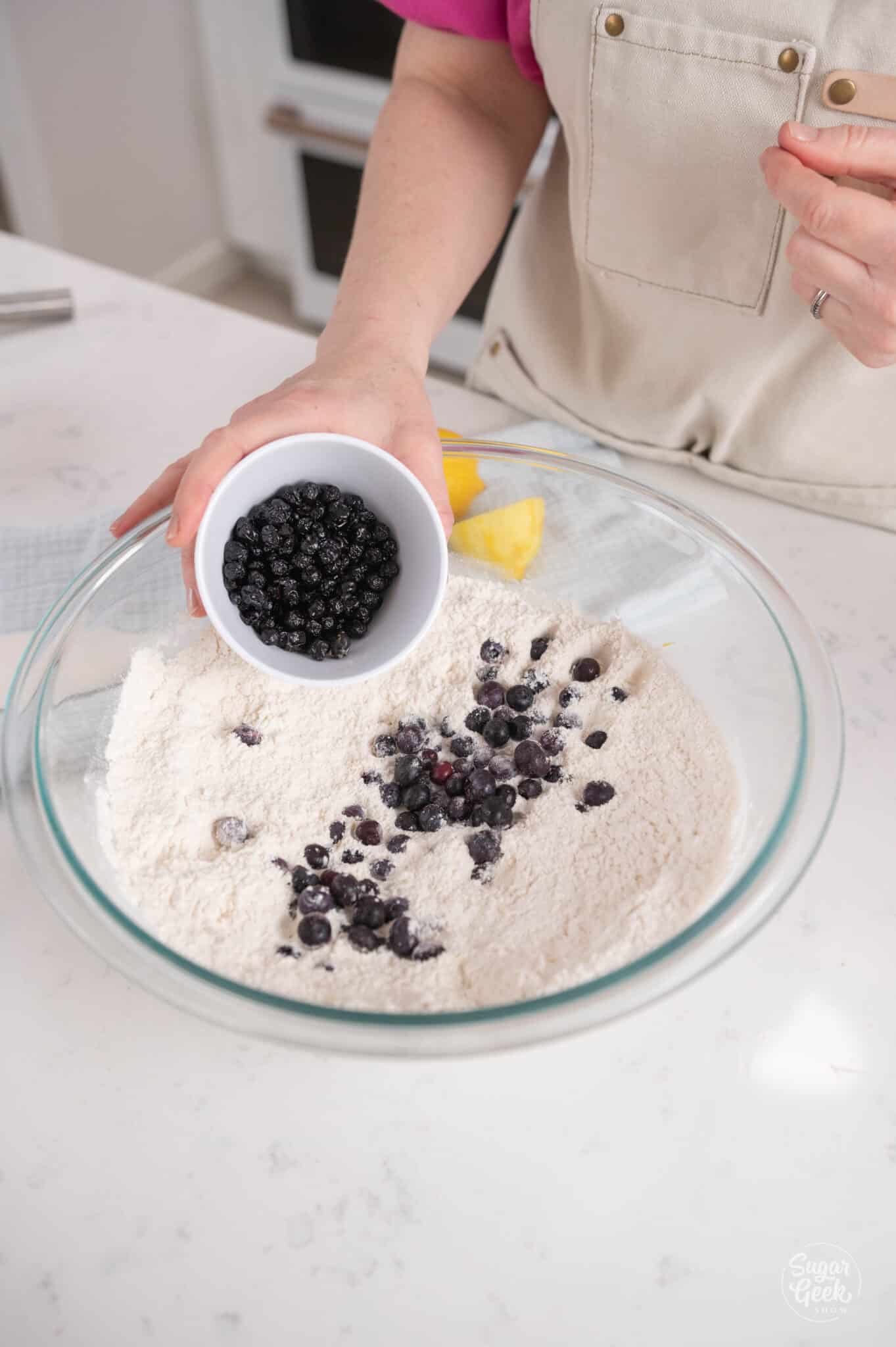 hand adding a cup of dried blueberries to a bowl of dry ingredients