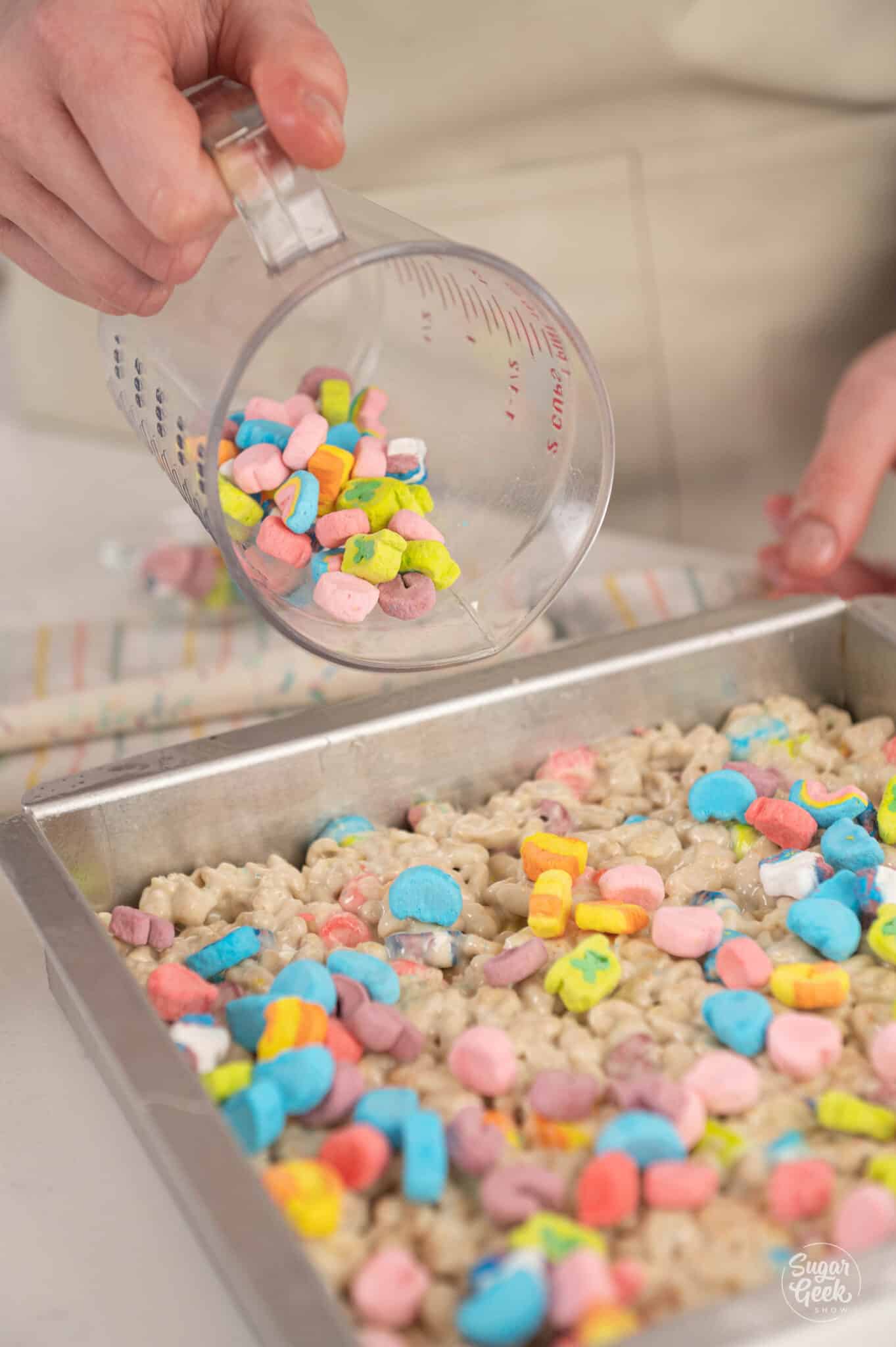 hand adding a container of lucky charm marshmallows on top of rice krispie treats
