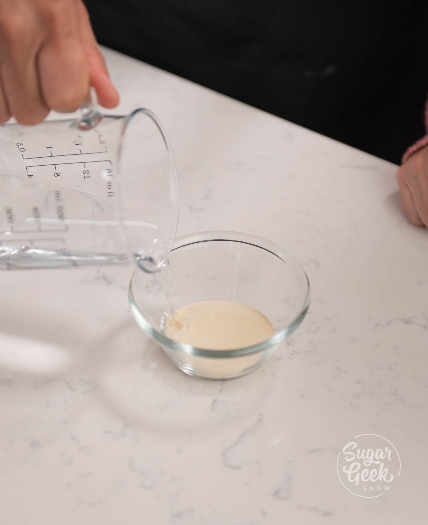 hand pouring measuring cup of water on to container of gelatin.