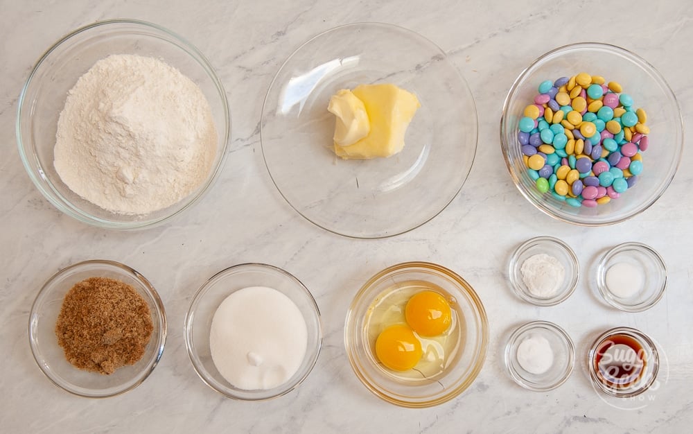 ingredients for M&M cookies in clear bowls on a white background shot from above
