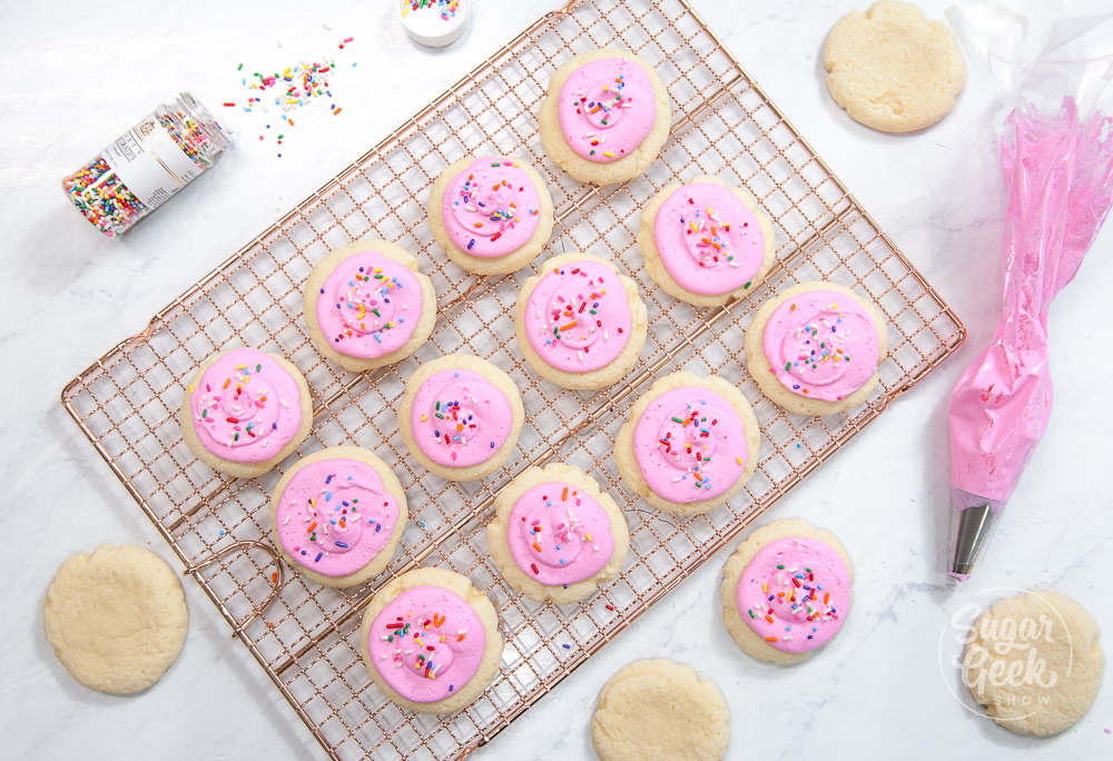 lofthouse cookies on a cooling rack with pink buttercream and sprinkles. Piping bag and sprinkles container off to the side