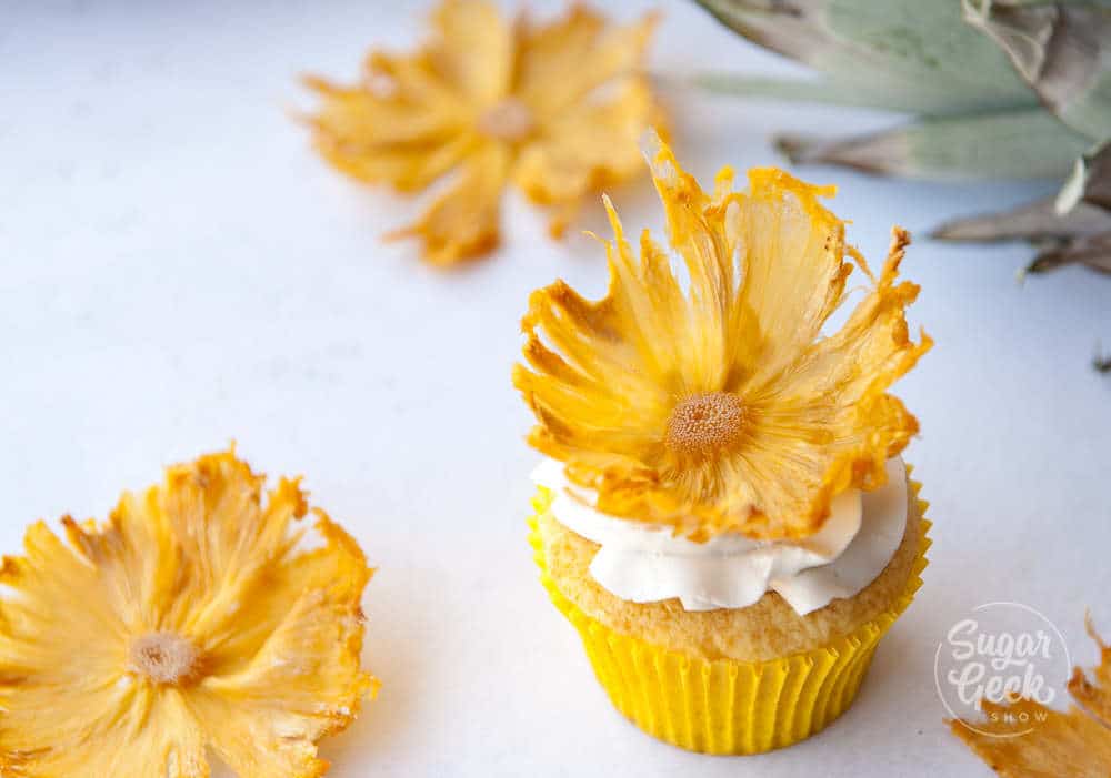dried pineapple flowers on vanilla cupcake with yellow wrapper and white buttercream frosting