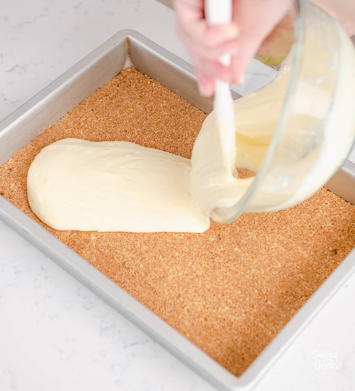 hands pouring a bowl of cheesecake batter over a graham cracker crust in a pan