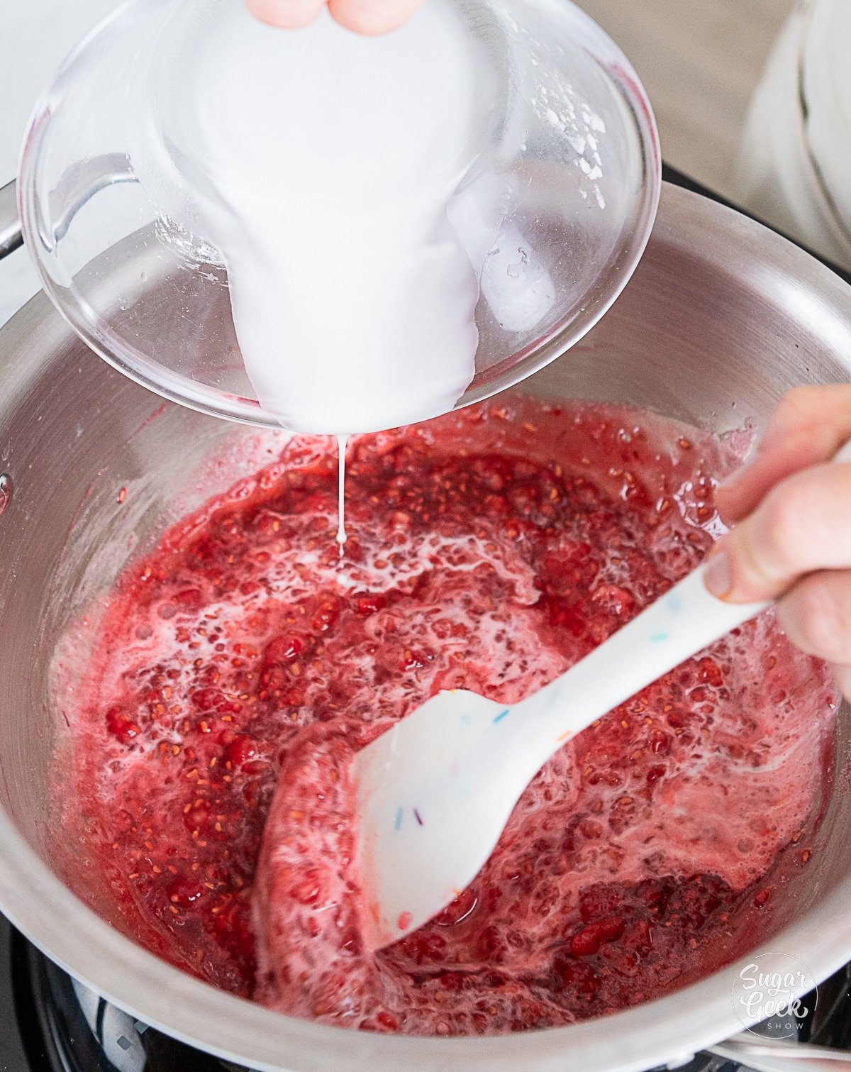 hand pouring cornstarch slurry into cooked raspberries in a pot while stirring.