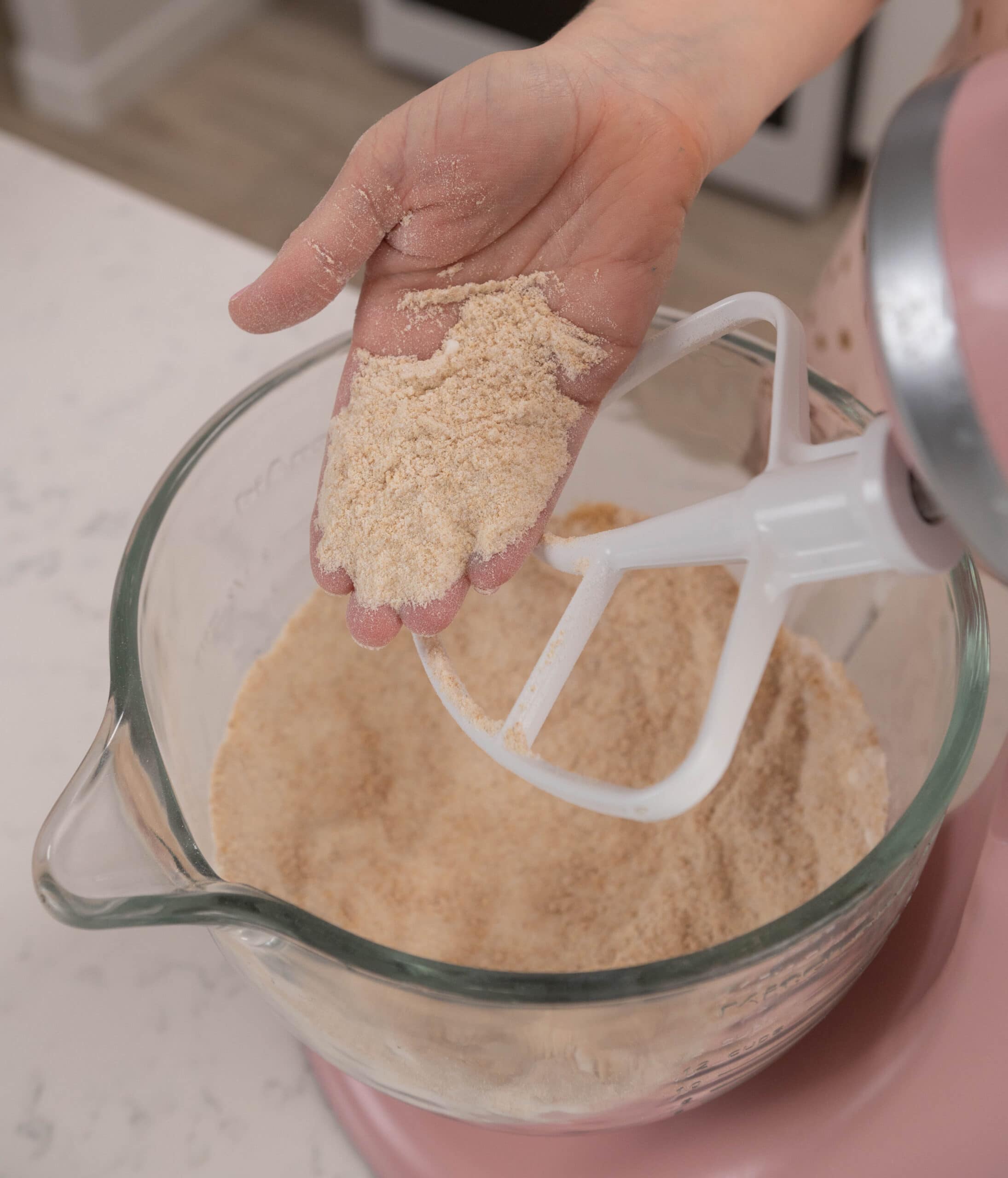 hand holding mixed dry ingredients above stand mixer bowl.