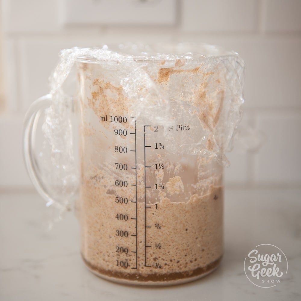 sourdough starter in clear measuring cup with plastic wrap on top