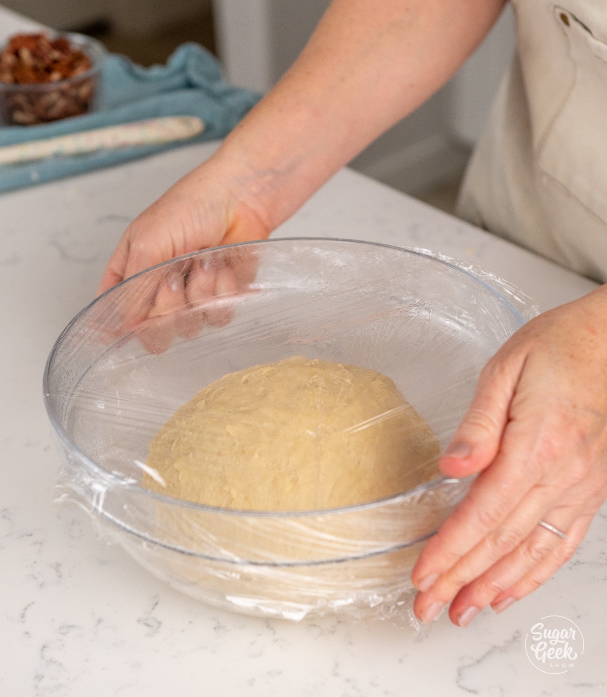 hands covering a ball of dough in a glass bowl with plastic wrap