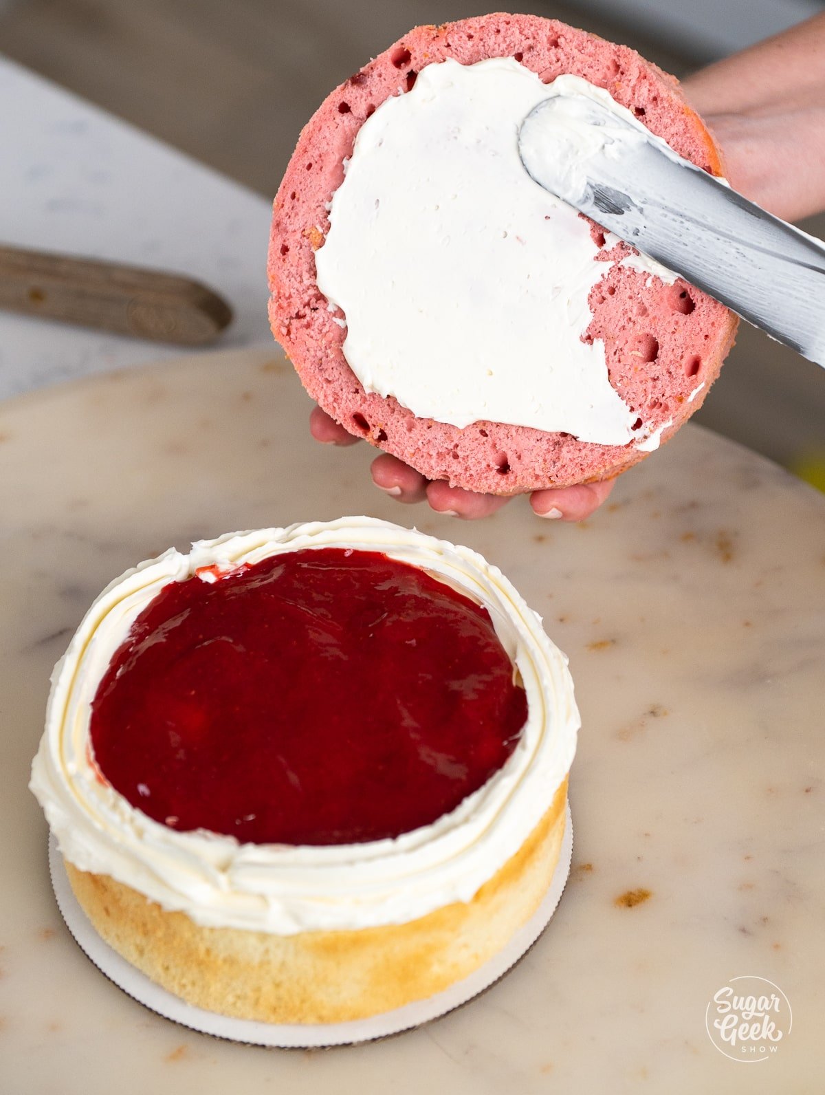 hands spreading buttercream onto the bottom of the strawberry layer of cake.