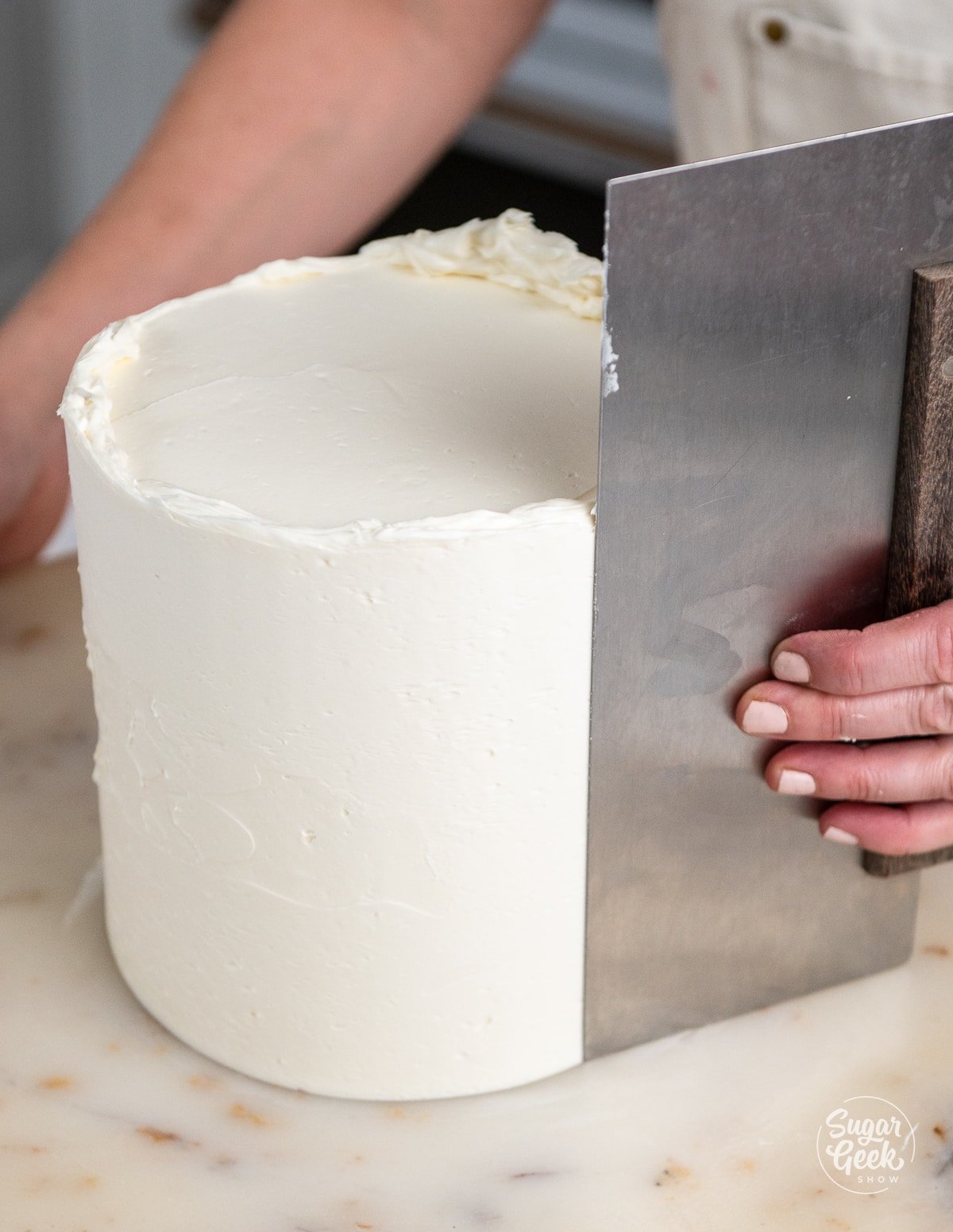 hands using a bench scraper to smooth the sides of the cake. 