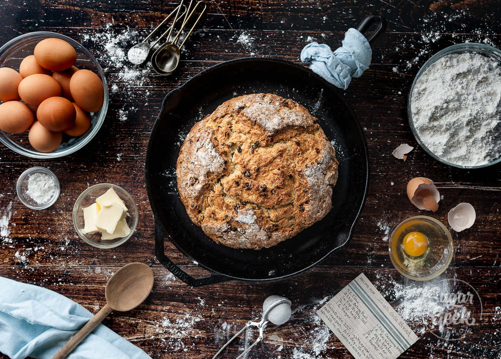 Irish soda bread in a cast iron pan surrounded by ingredients on a wooden backdrop