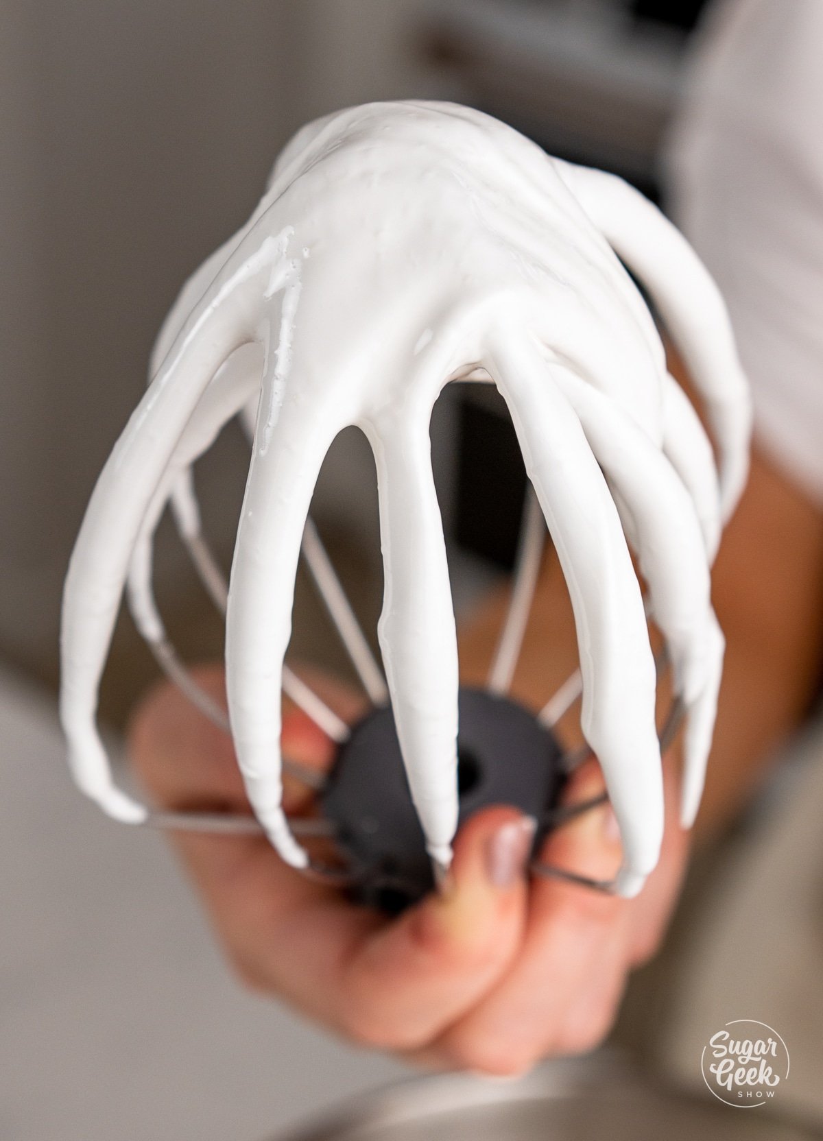 hand showing whisk attachment with glossy stiff peaks. 